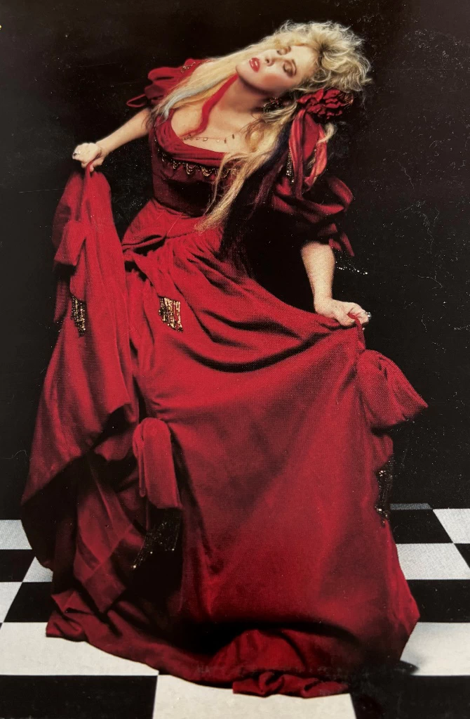 Stevie Nicks in an elaborate red dress flourishes in a dance on a checkerboard floor.